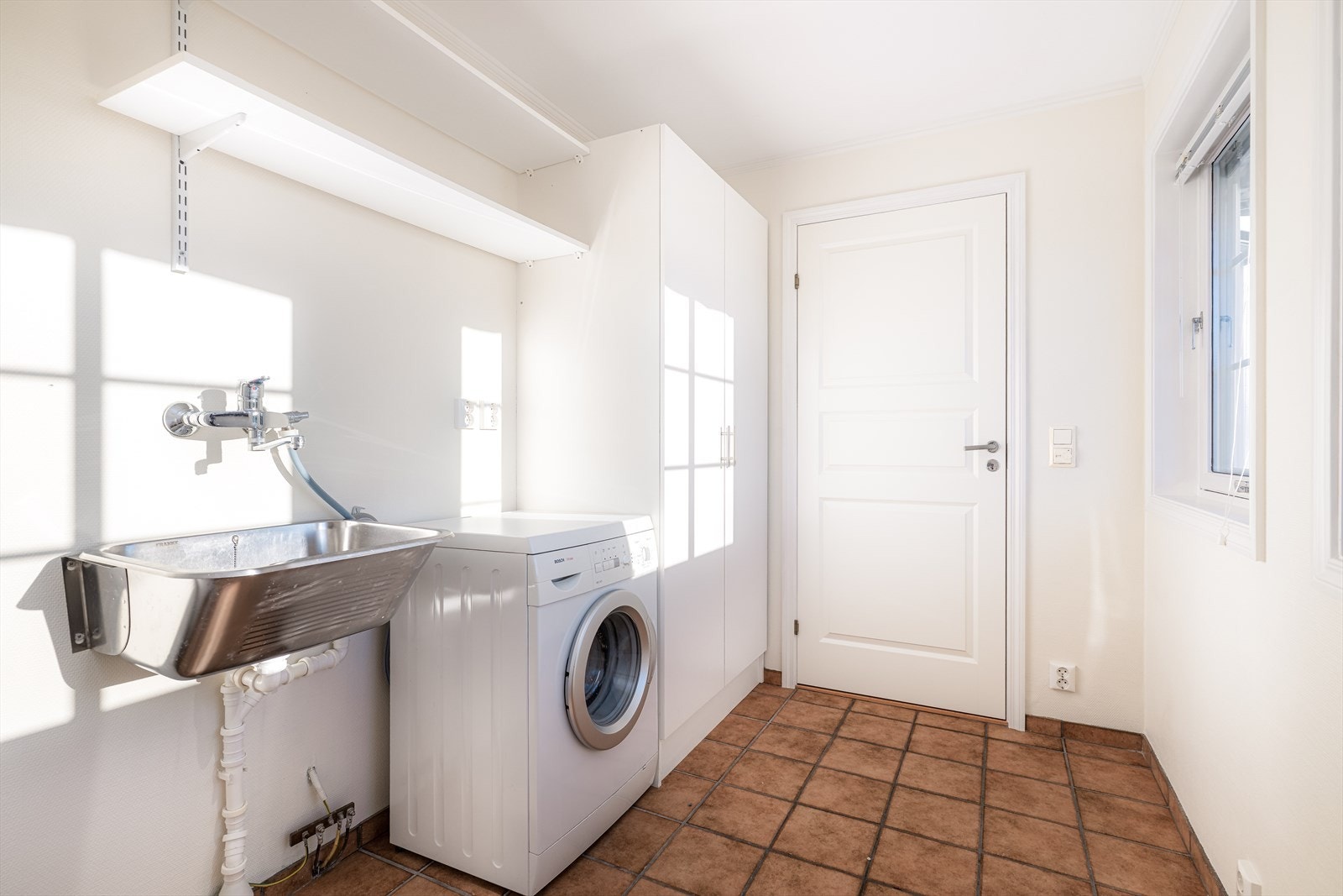 Vaskerom med flis og varmekabler på gulv, og ellers malte overflater. Vaskerommet er innredet med skap, stålkum og opplegg for vaskemaskin. Galleribilde