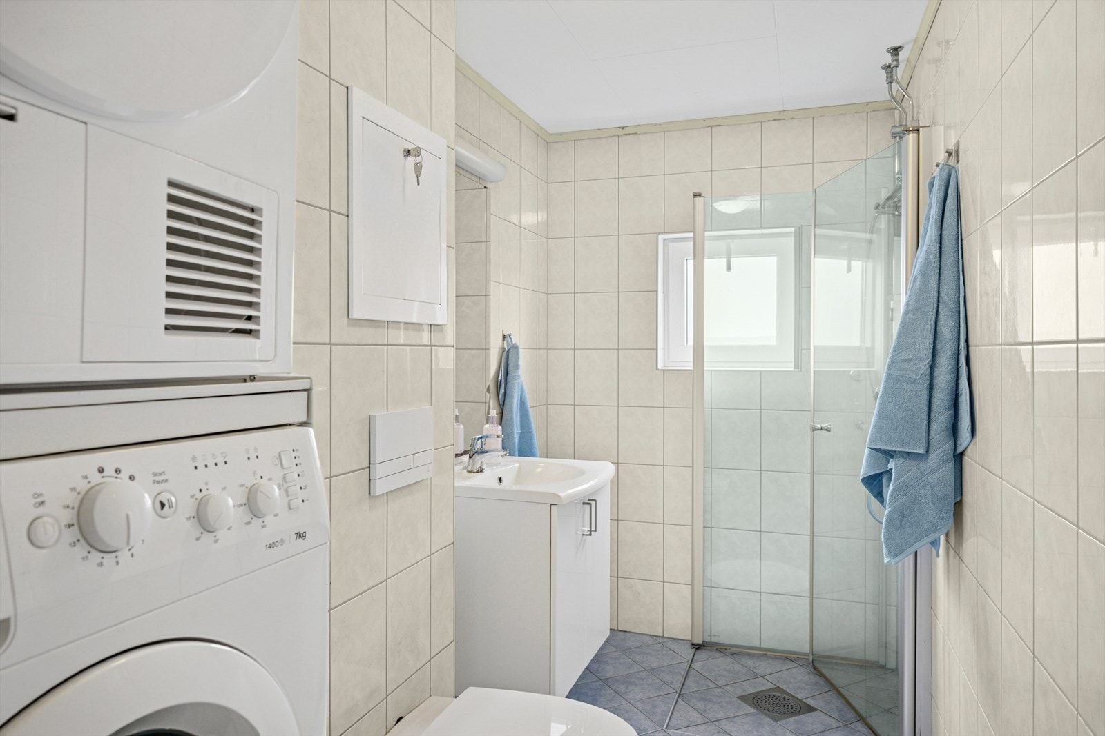 Flislagt bad som inneholder servant nedfelt i skuffeseksjon, toalett, dusj i hjørne og opplegg for vaskemaskin. Galleribilde