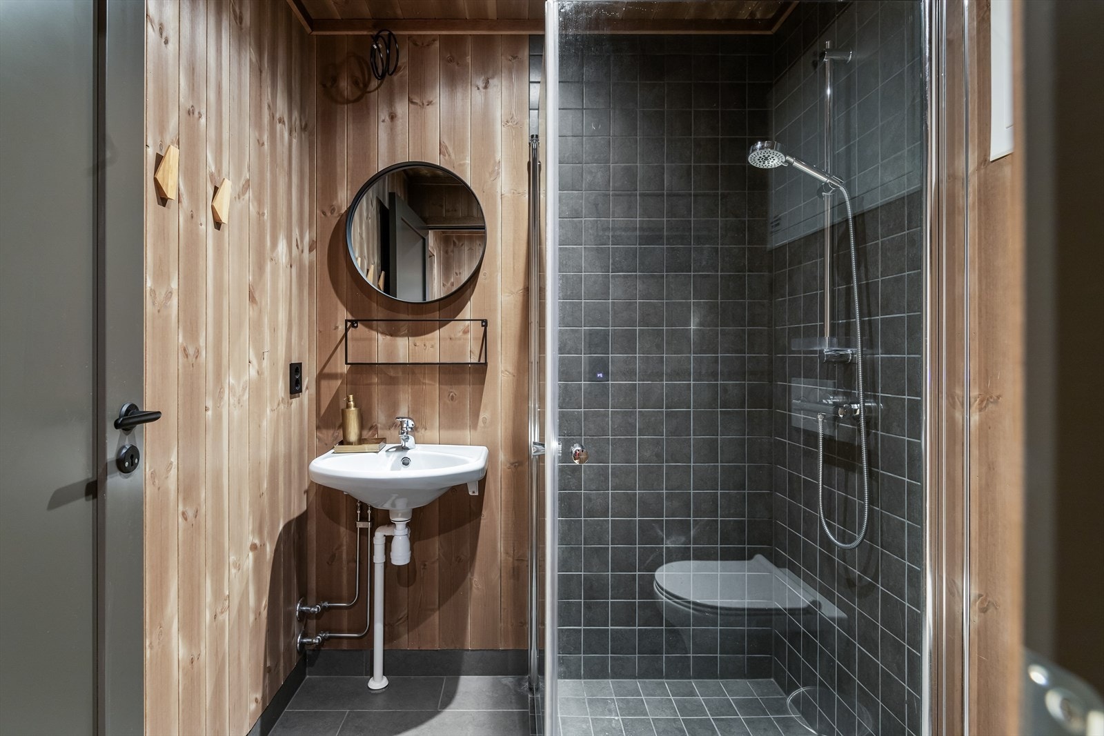 På gulvene, lang sokler og vegger i dusjhjørner er det grå fliser i to ulike størrelser. Hvert dusjhjørnet er også utstyrt med veggmontert hånddusj og rette, innfellbare glassdører. Videre har badene downlights, gulvvarme og vegghengte toalett. Galleribilde