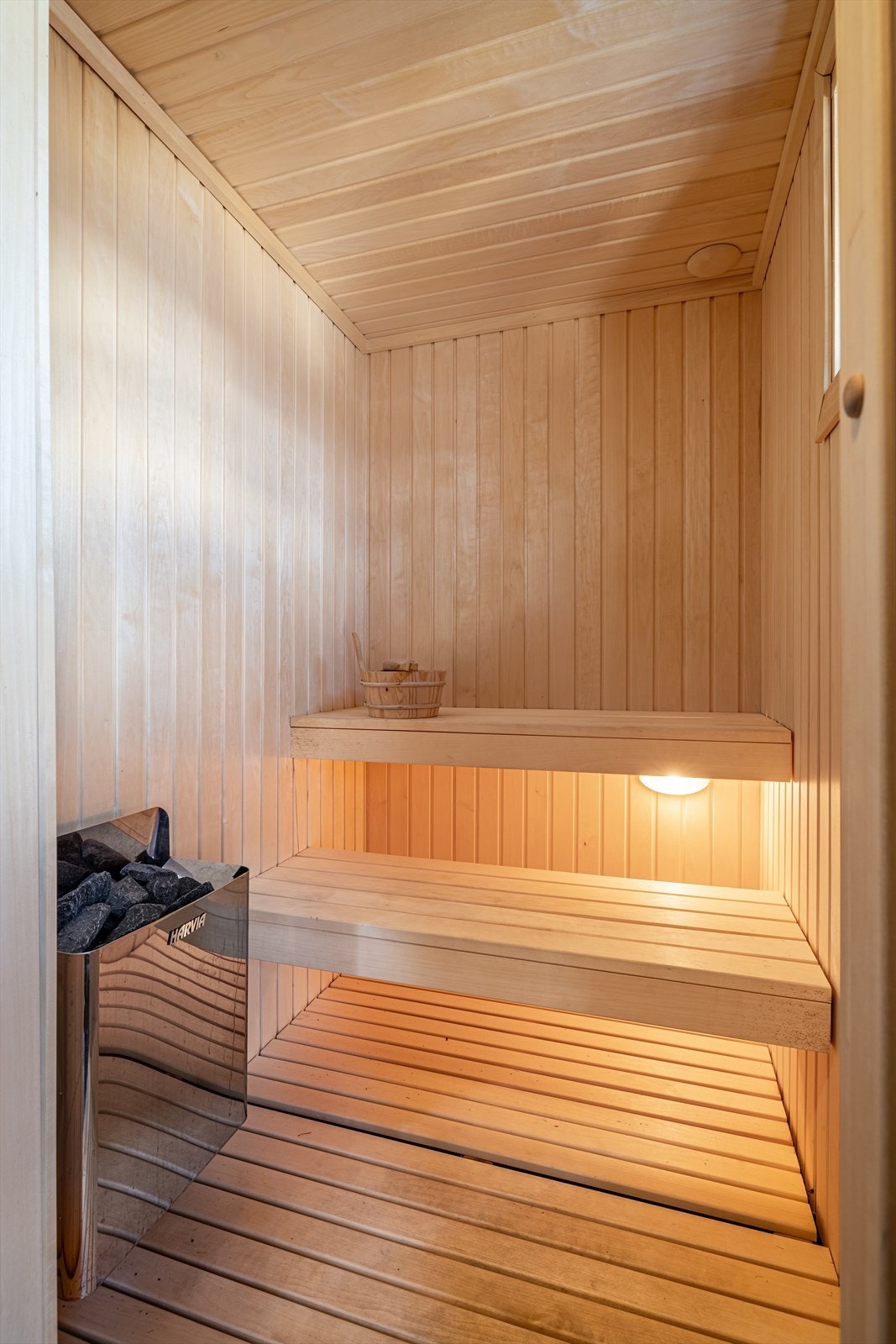 Det er en badstue for høy komfort, innredet med tremmegulv og trepanel på veggene. En elektrisk badstueovn er installert, og rommet har glassdør og plassbygde trebenker med belysning under. Galleribilde