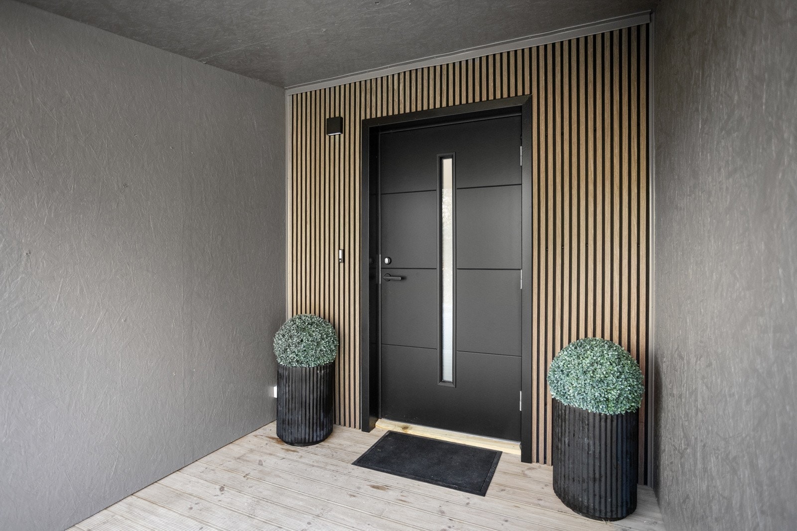 Inngangspartiet er overbygget, har belysning og spiler på vegg og moderne inngangsdør med glassfelt. Galleribilde