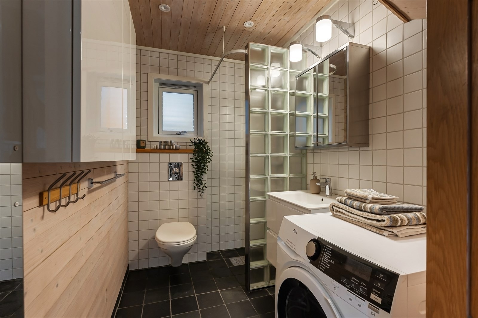 Baderom fra byggeår med dusjhjørne, veggfestet toalett, benkeskap med servant og overskap med speil samt opplegg for vaskemaskin Galleribilde