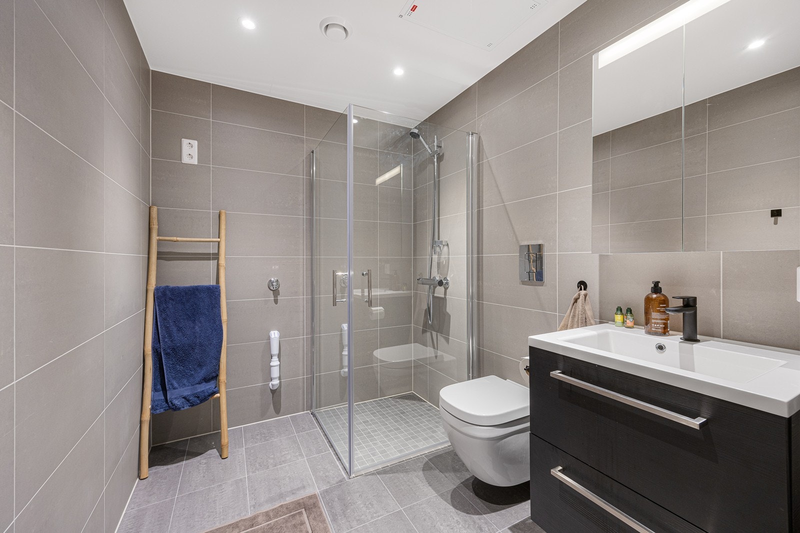 Badet er av god størrelse og har fliser i en lun gråfarge. Det er gulvvarme, vegghengt toalett, dushjørne med glassdører og det er opplegg for vaskemaskin Galleribilde