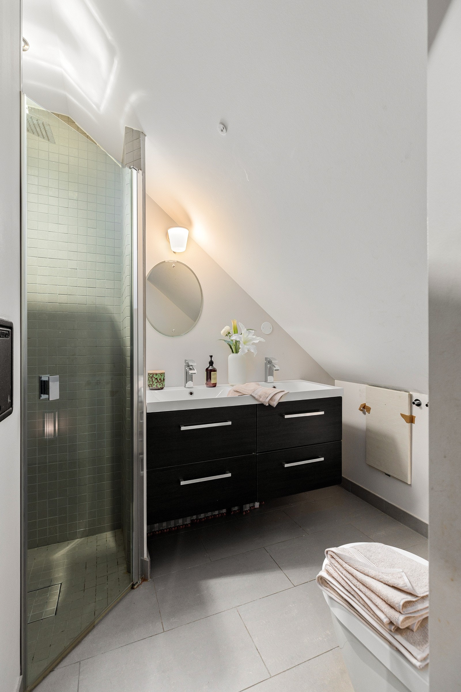 Badet på loftet er innredet med flislagt gulv og dusjnisje, dobbelservant, mørk skuffeseksjon og veggmontert toalett.