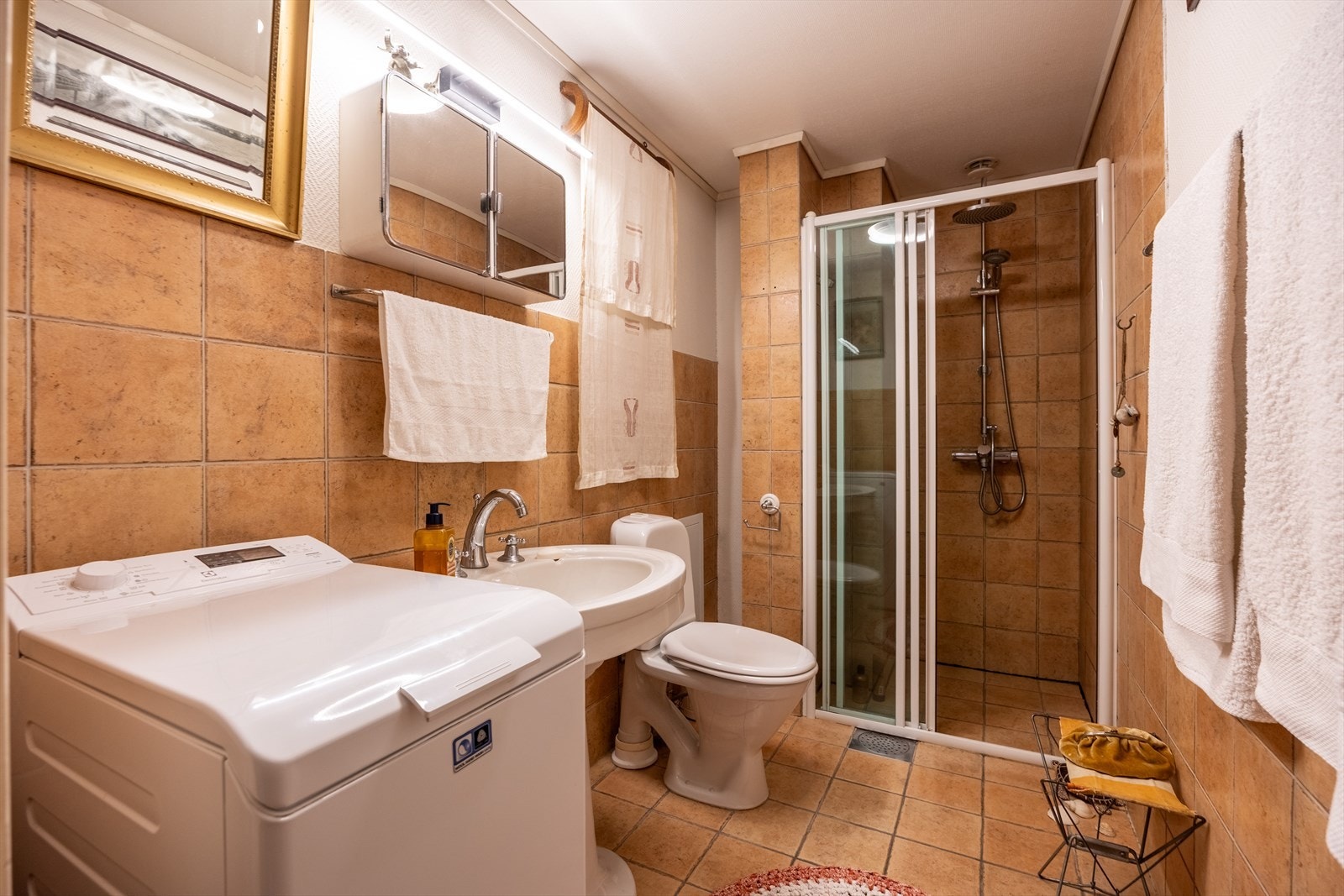 Flislagt eldre bad med gulvvarme utstyrt med dusjhjørne, innfellbare dusjdører, naturlig avtrekk, servant, servantskap, opplegg for vaskemaskin, rørfordelingsskap og wc Galleribilde