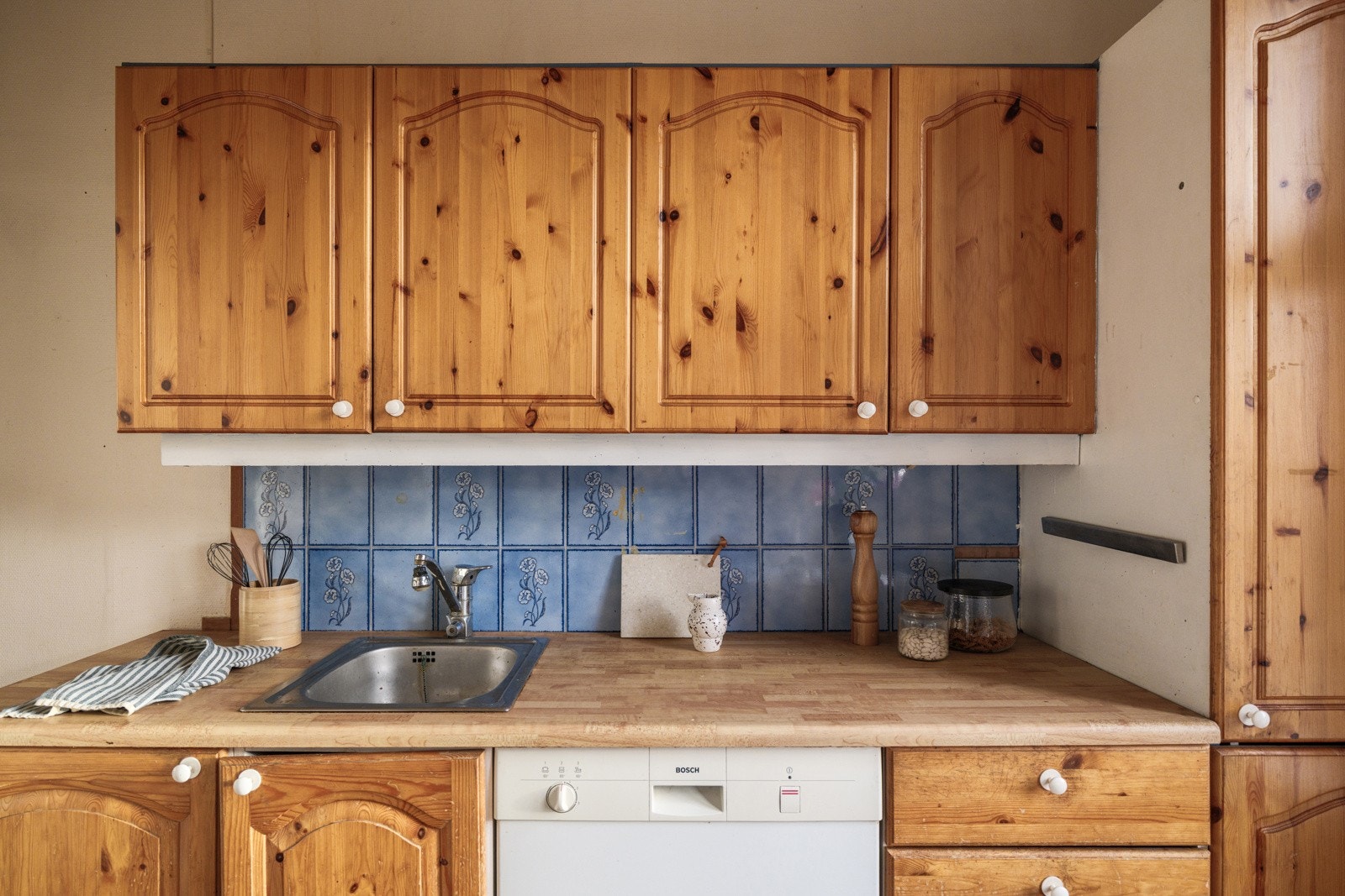 Eldre utdatert kjøkkeninnredning med profilerte furu fronter, laminert benkeplate med nedfelt oppvaskkum og fliser i benkeryggen. Galleribilde