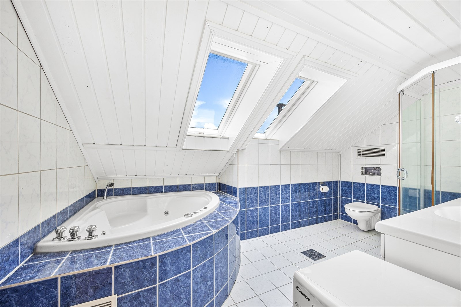 Flislagt bad med takvinduer og innredning bestående av klosett, badekar, dusjkabinett og baderomsinnredning med overskap og speil Galleribilde