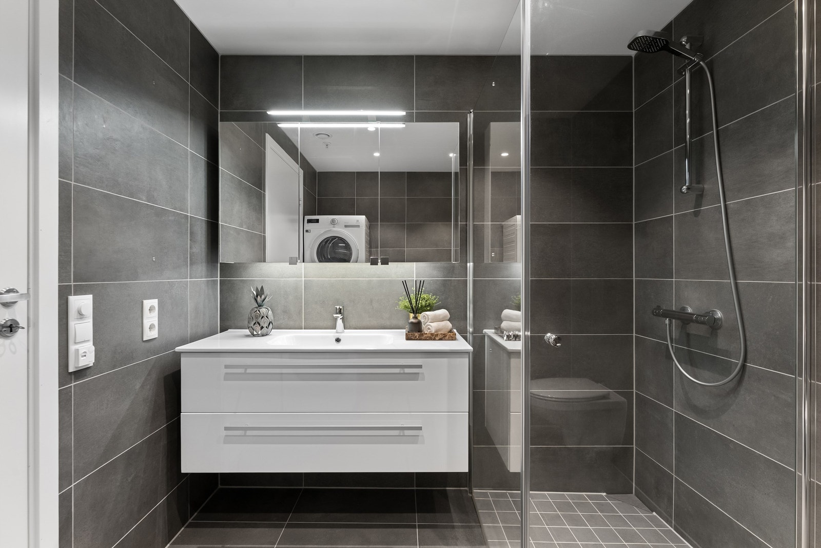 Badet har vegghengt toalett, servant, speil, dusjhjørne og opplegg for vaskemaskin Galleribilde