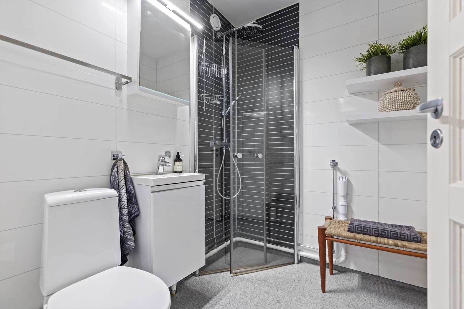 Badet er innredet med gulvstående toalett, heldekkende servant med skap under, overskap med speilfront og lys, dusjhjørne med falldusj og dører i herdet glass. Det er opplegg for vaskemaskin. Galleribilde