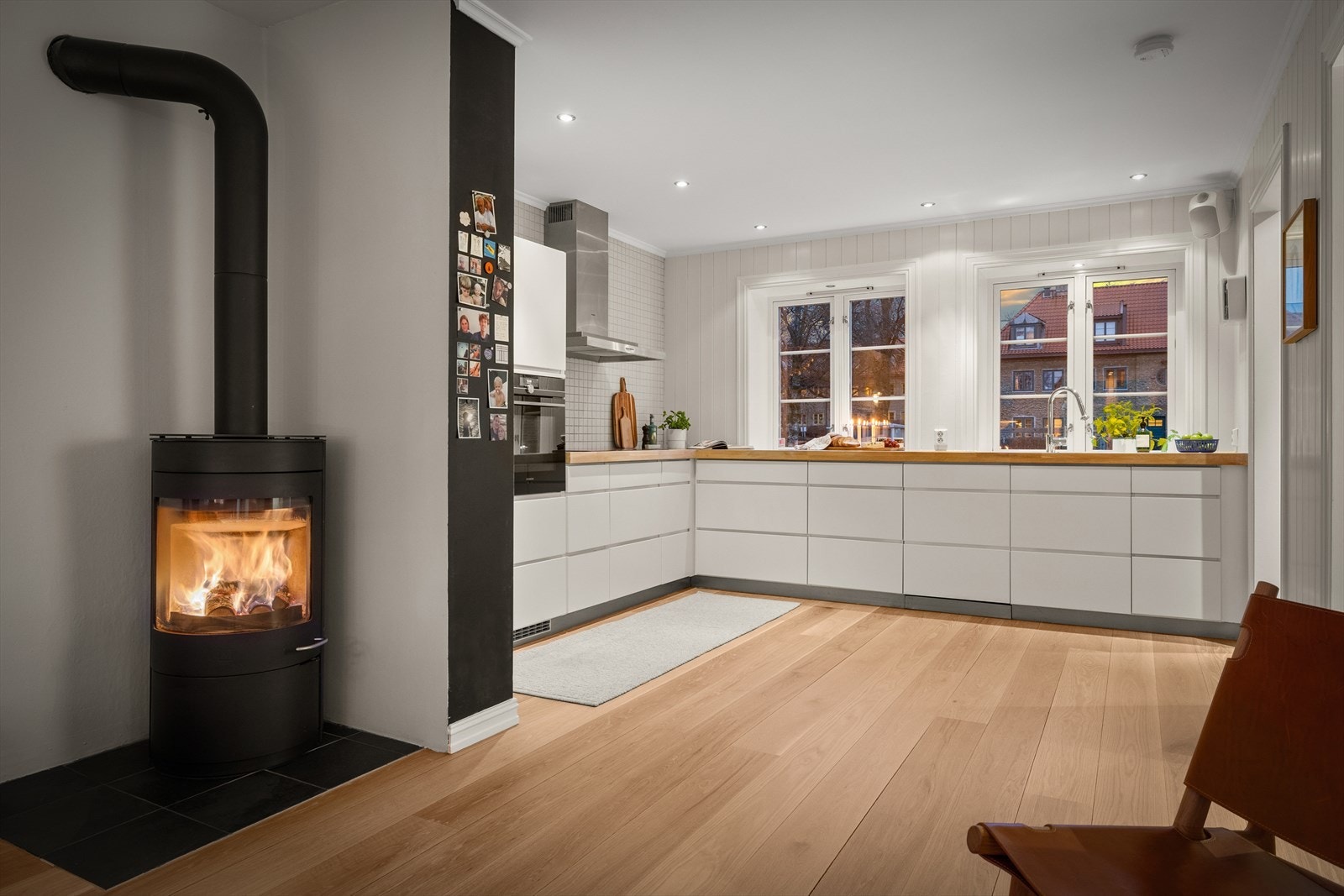 Vinduer mot de grønne omgivelsene gir subtilt kveldsskjær utenfra, og det åpne samspillet med spisestuen gjør at gløden fra den vedfyrte peisen kan skimtes inn mot kjøkkenet. Galleribilde