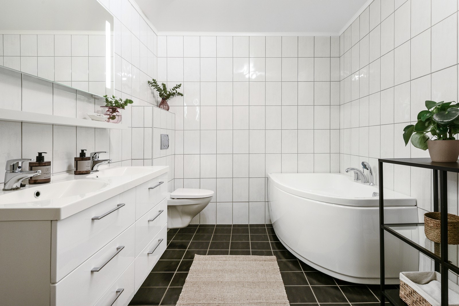 Badet i underetasjen følger samme stil som badet i hovedetasjen, men er romsligere. Det har stort hjørnebadekar med dusjmulighet, vegghengt toalett og dobbelt servant med oppbevaring. Galleribilde