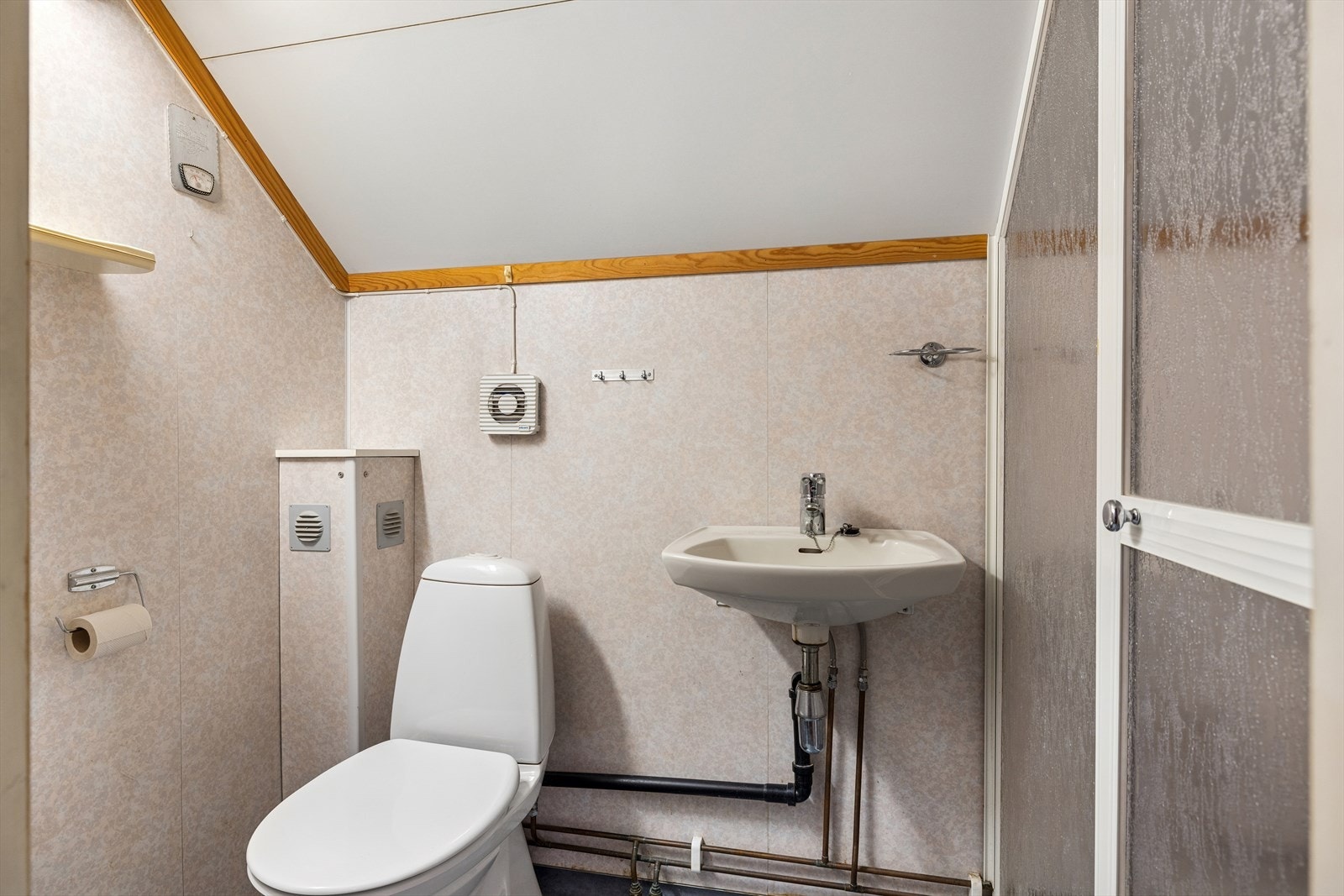 Badet er fra før 1997 og har baderomsplater på veggene, belegg på gulvet og malte plater i taket. Rommet er innredet med en vegghengt servant med ettgreps blandebatteri, et gulvmontert toalett og en dusjnisje med skyvedør. Galleribilde