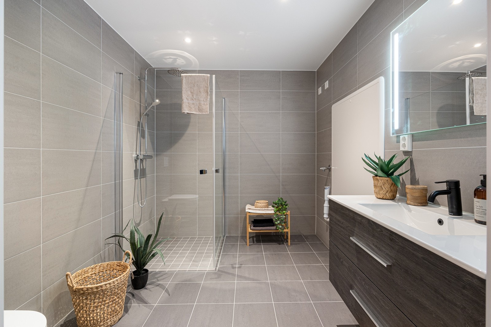 Badet er delikat utformet med grå fliser på gulv og vegger, gulvvarme og downlights. Dusjhjørne i aluminium og glass med påkostet regnfallsdusj og hånddusj på stang Galleribilde