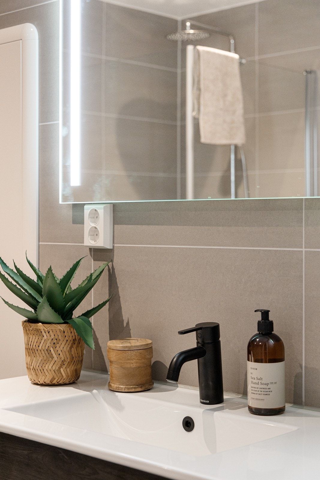 Baderomsinnredning med skuffer i mørk utførelse, heldekkende servant, ett-greps batteri i sort utførelse og tilhørende speil med lys Galleribilde