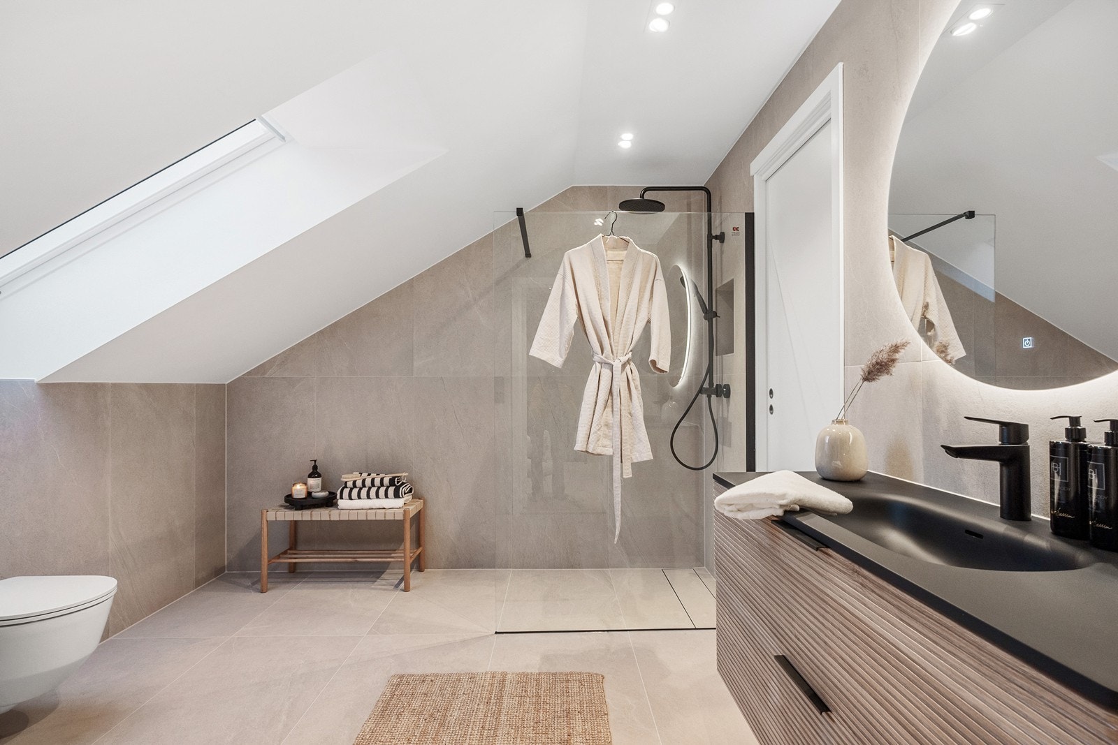 Badet matcher boligens stil med lyse 60x120 fliser, sort servant med tilhørende speil og vegghengt toalett. Dusjhjørnet er adskilt med glassvegg, har innebygd nisje, samt hånd- og takdusj. Galleribilde