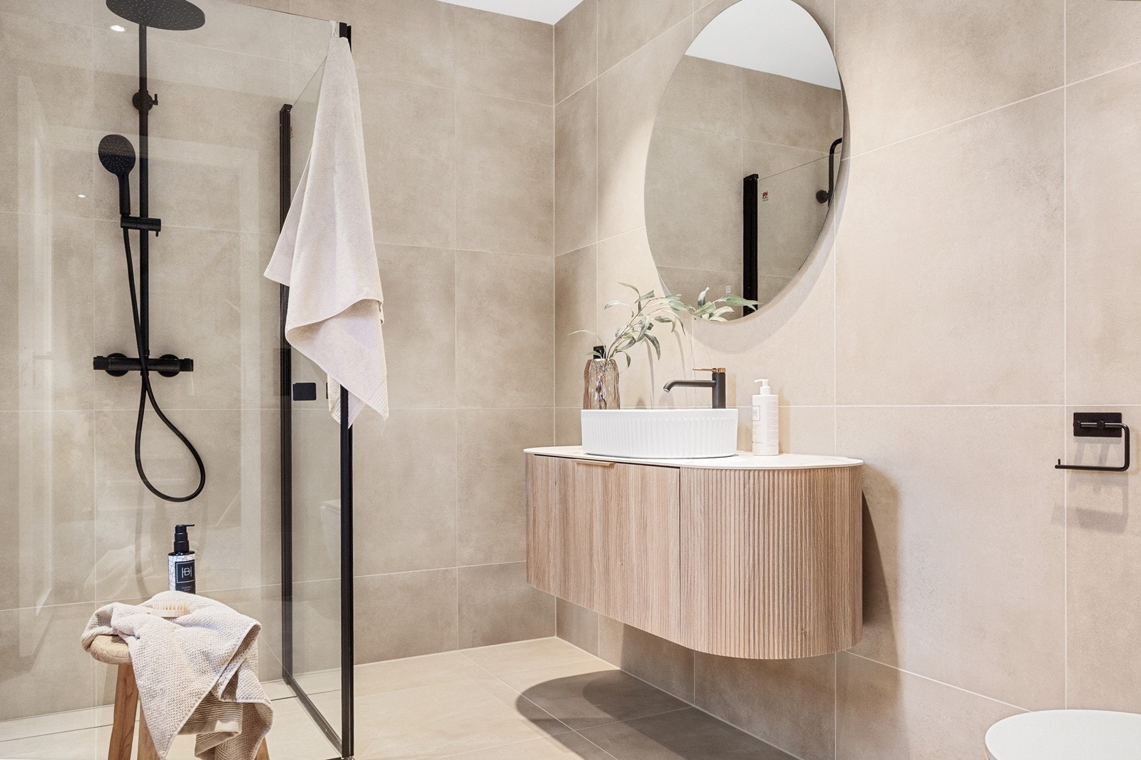 Badet i første etasje har lyse 60x60-fliser, vegghengt toalett og servant i eik med tilhørende bolleservant. Det runde speilet og sorte armaturene gir et moderne uttrykk, mens dusjsonen med innfellbare glassdører og både hånd- og regndusj. Galleribilde