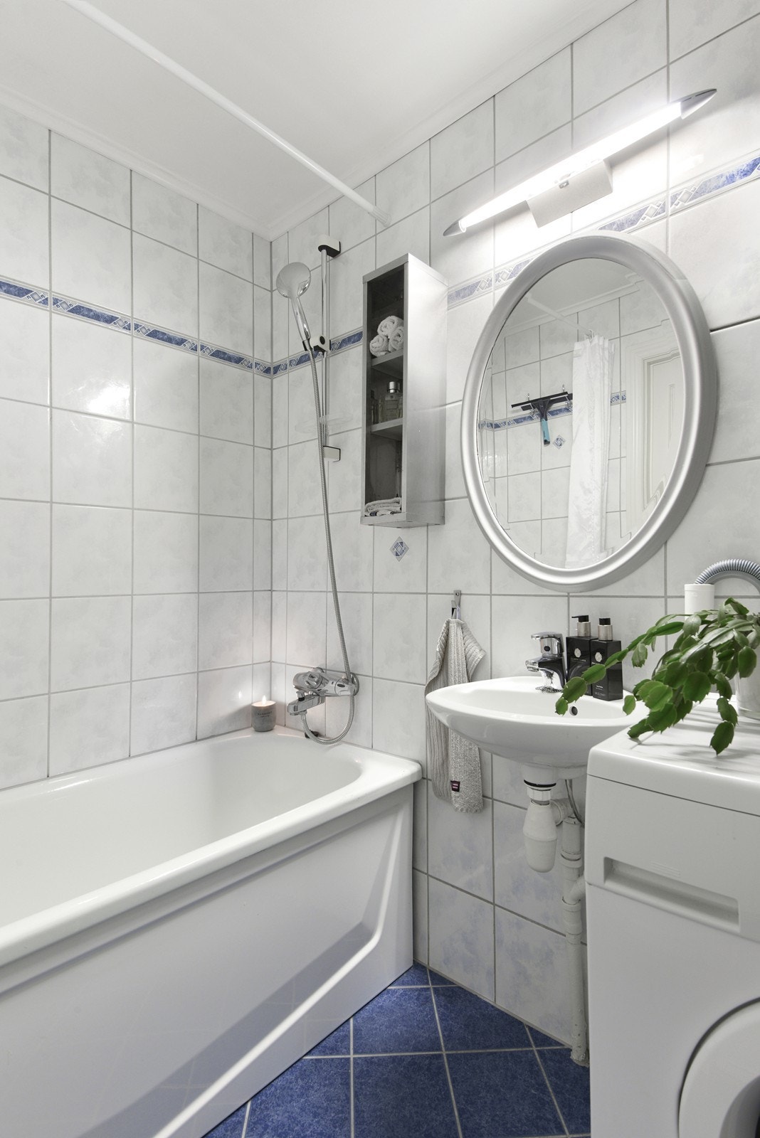 Badet har ukjent alder og flislagte overflater i hvite og blå toner. Rommet ligger rett ved gangen og er innredet med badekar med dusjmulighet, gulvfestet toalett og vask med speil og belysning. Galleribilde