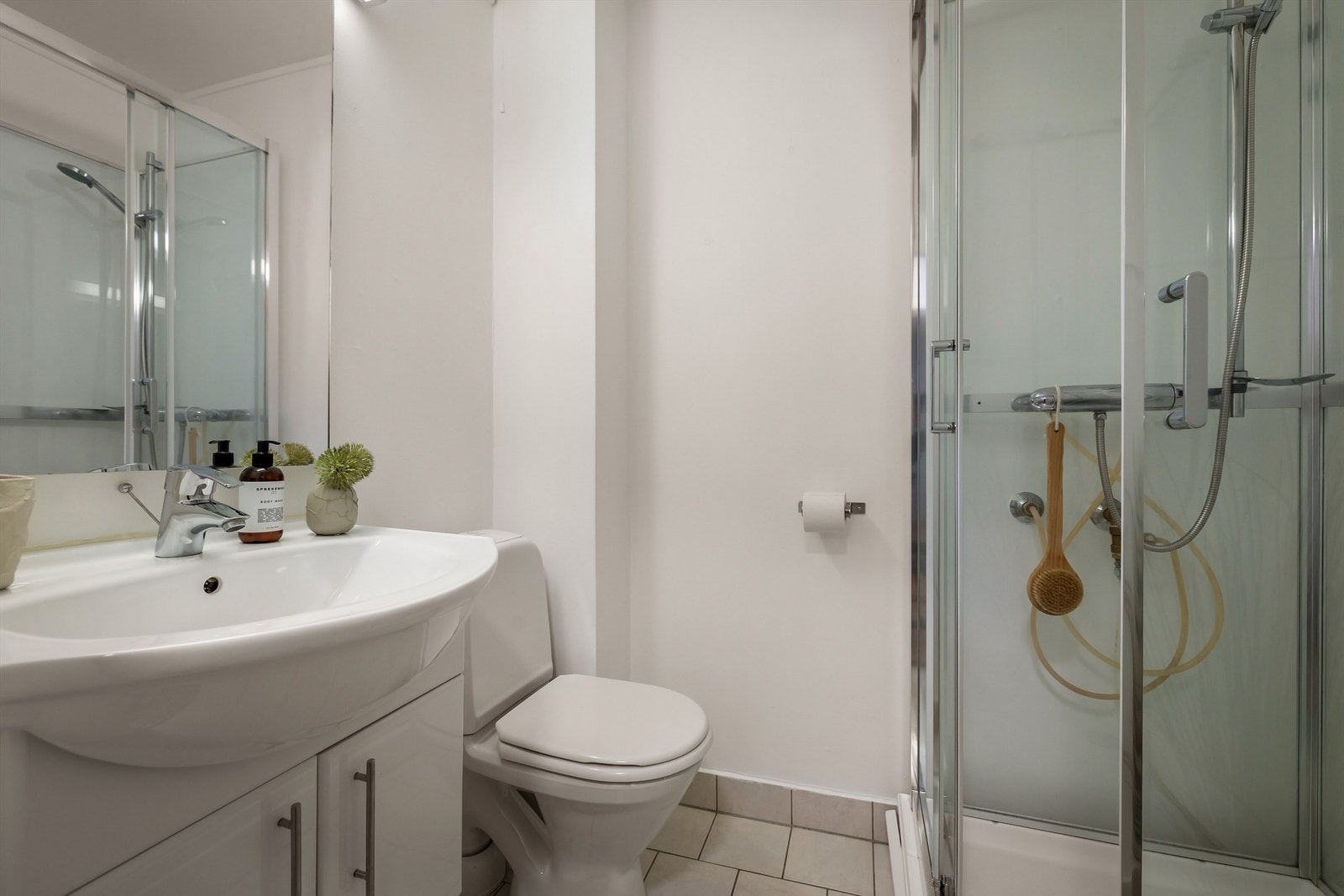 Flislagt baderom innredet med gulvstående toalett, servant med skapinnredning og speil med belysning over Galleribilde