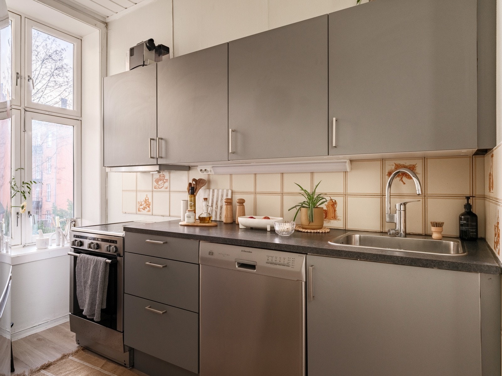 Kjøkkeninnredning med grå glatte fronter og laminert benkeplate med nedfelt oppvaskkum. Det er fliser i benkeryggen og belysning under overskap. Galleribilde