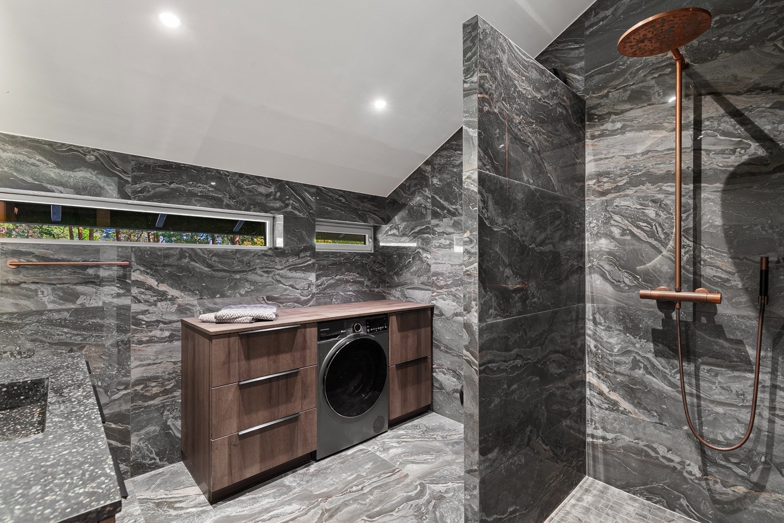 Videre er badet utstyrt med servant med skuffer, vegghengt toalett og dusjhjørne med tak- og hånddusj i kobber, samt opplegg for vaskemaskin. Vask og benkeplate fra Øvereng Interiør. Galleribilde