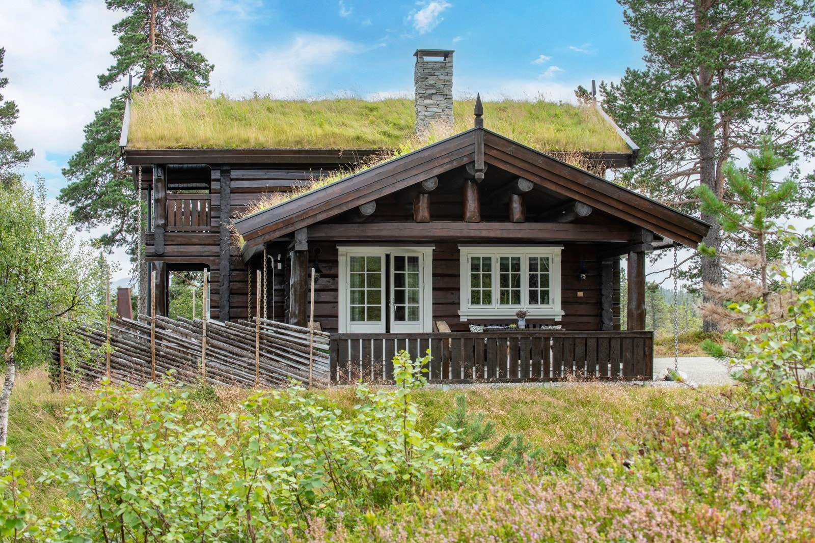 Tradisjonell tømmerhytte med torvtak, stor veranda, omgitt av frodig natur. Galleribilde