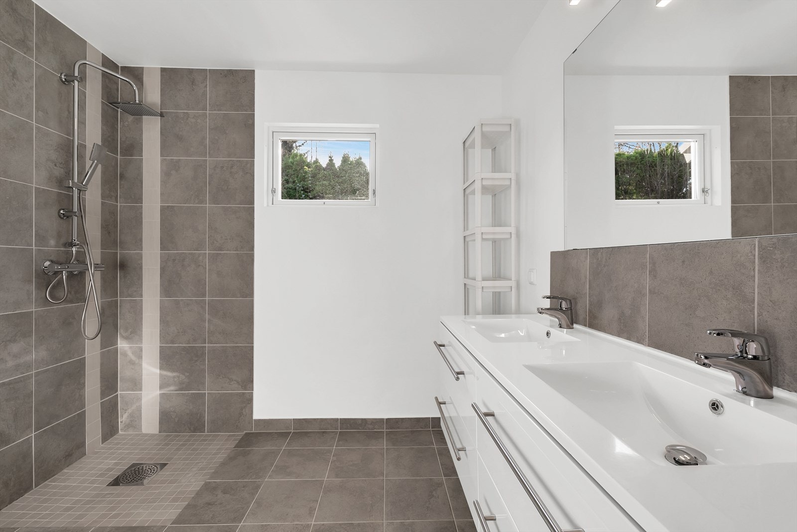 Lekkert baderom fra 2024 med innredning som består av benkeskap med glatte fronter, to praktiske servant, veggfestet klosett med innebygget sisterne og dusjhjørne med dører i herdet glass. Galleribilde