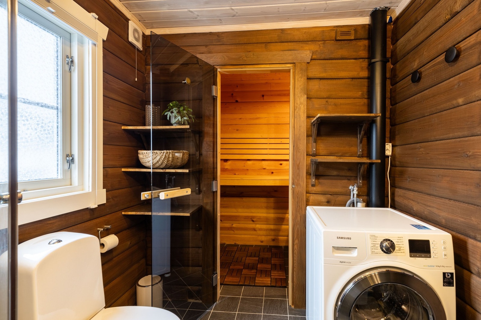 Badet har glassdør inn til en badstue med flislagt gulv, badstueovn og plassbygde trebenker. Galleribilde