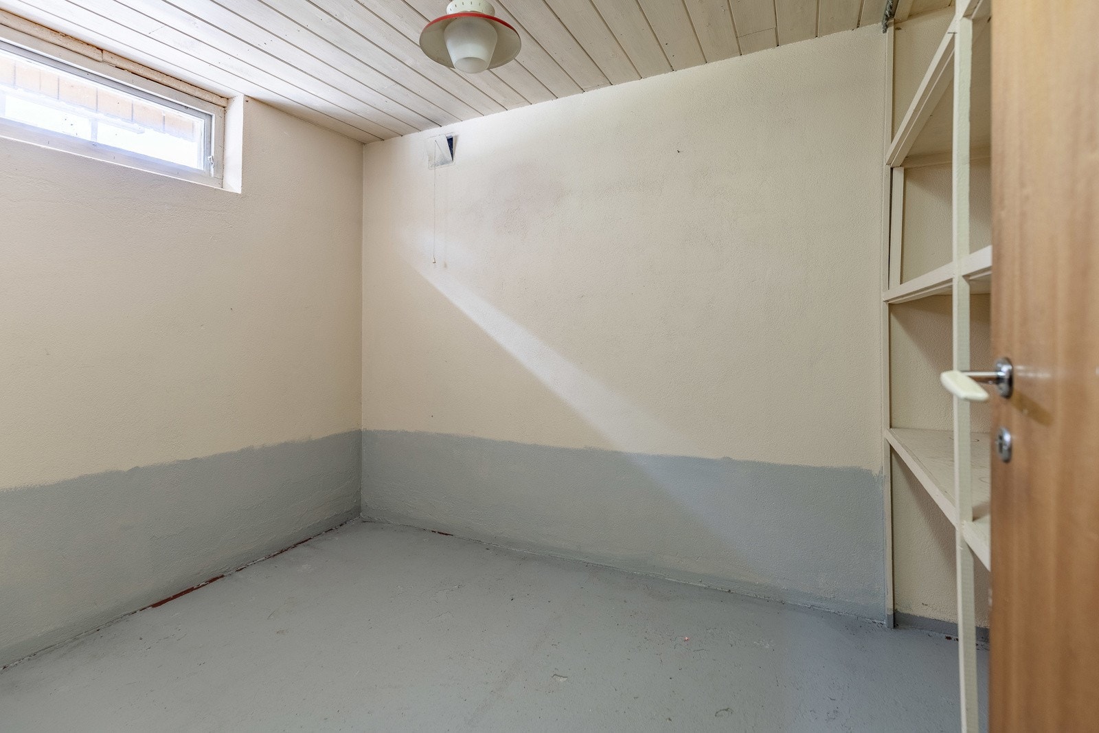 Rommet har grått betonggulv, beige vegger med grå nederst, og et lite vindu. Galleribilde