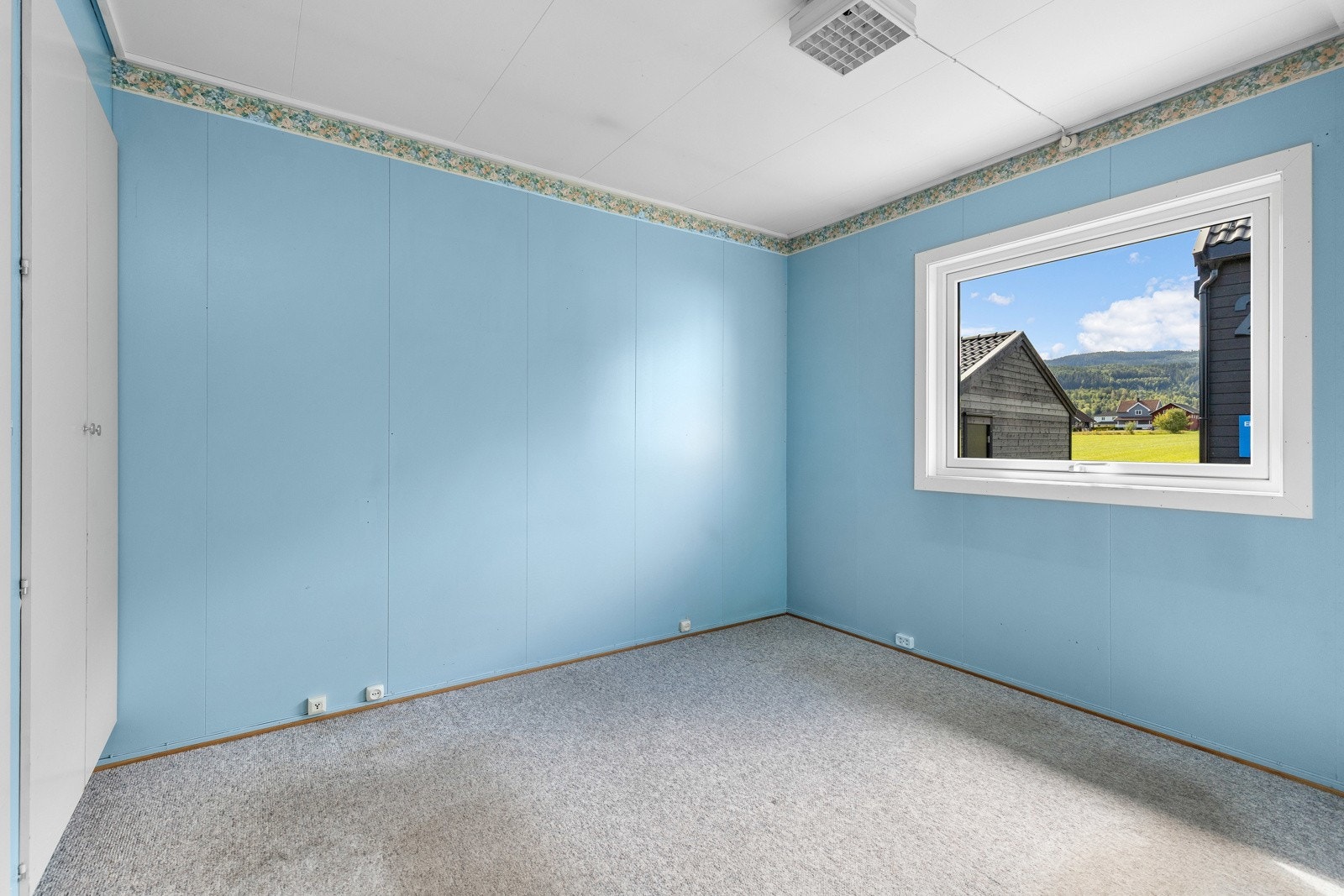 Rommet har lyseblå vegger, grått gulvteppe og et stort vindu som gir utsikt til nabolaget. Galleribilde