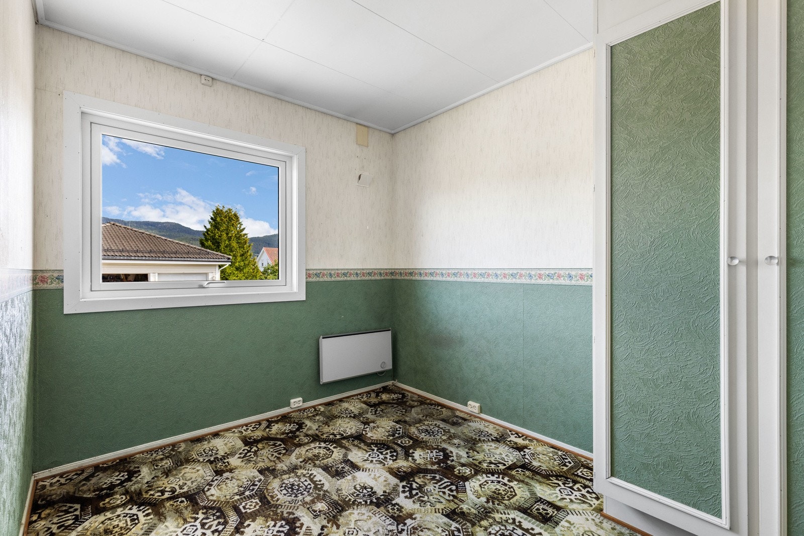 Rommet har tapet på veggene, teppe på gulvet, elektrisk ovn og vindu med utsikt. Galleribilde