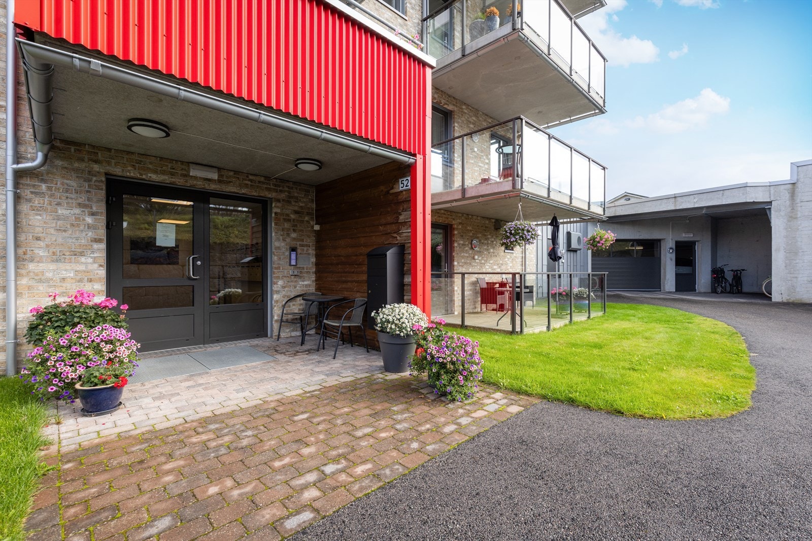 Leilighetsbygget har felles, overbygget inngangsparti på bakkeplan, med belegningsstein og utebelysning, samt ringetablå. Til høyere i bildet ser du inngang til felles garasjeanlegg, hvor leiligheten disponerer bod og en biloppstillingsplass. Galleribilde