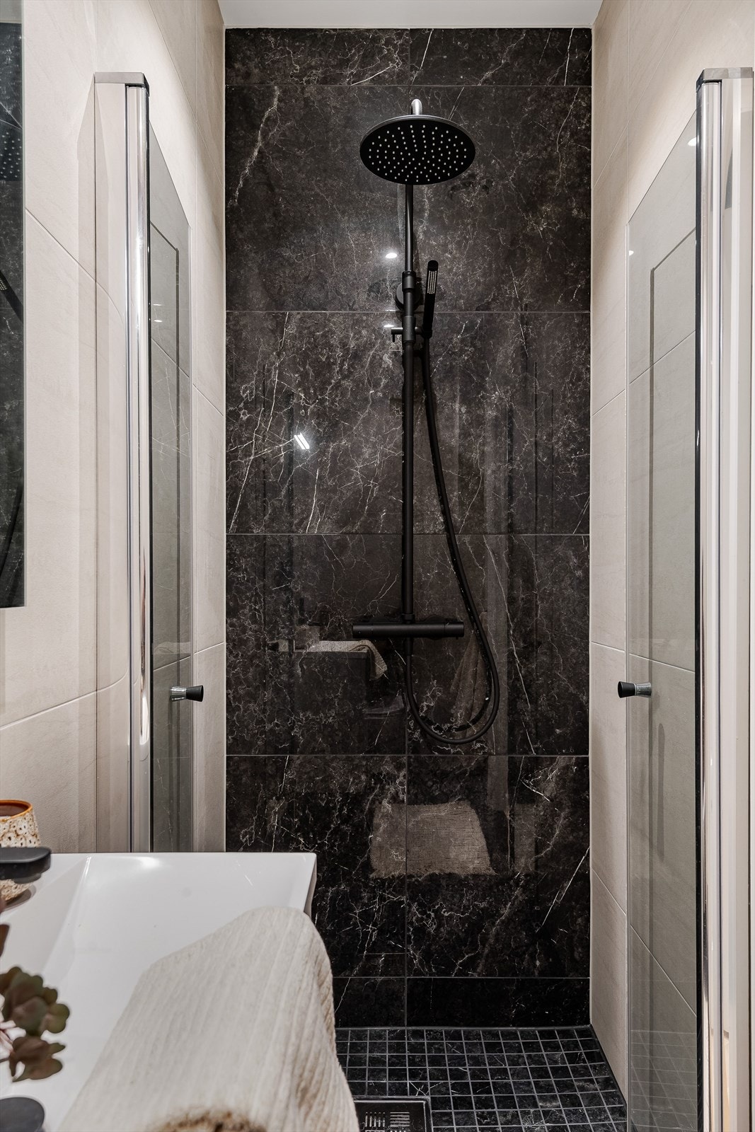 Dusjnisje med innfellbare dusjdører og veggmontert dusjbatteri med regnfall. Mørk flis i dusjområdet skaper en elegant kontrast til de lysere veggene. Galleribilde