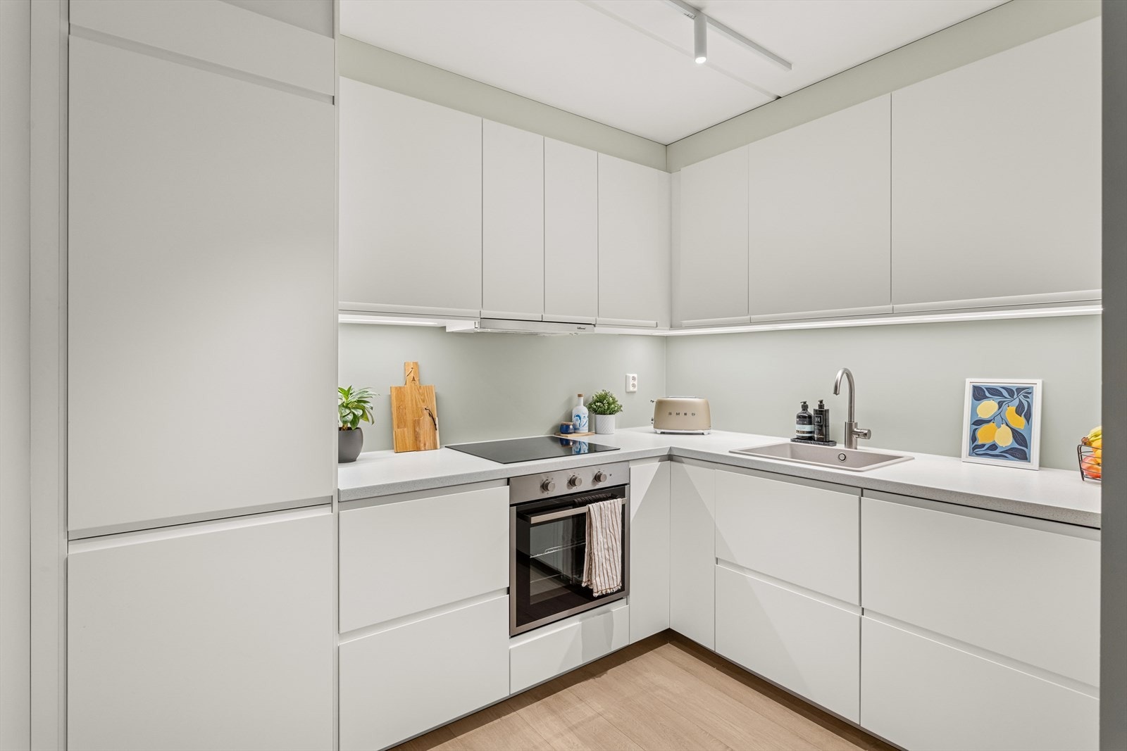 Pen Ikea kjøkkeninnredning fra 2023 med glatte fronter og en benkeplate i laminat. Hvitevarer som platetopp (2021), stekeovn (2021), oppvaskmaskin (2023) og kjøle/fryseskap (2023) er integrert i innredningen. Galleribilde