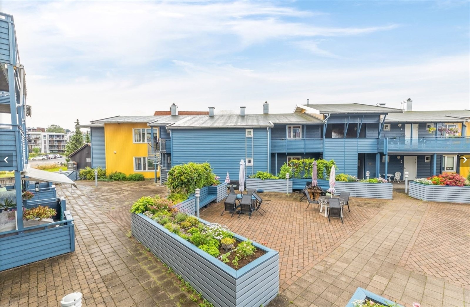 Bilde fra tidligere anledning som viser at tomten er pent opparbeidet med grøntareal, busker og trær, samt hyggelige uteplasser med belegningsstein og blomsterkasser med beplantning. Her er det bord og stoler til felles bruk og hygge Galleribilde