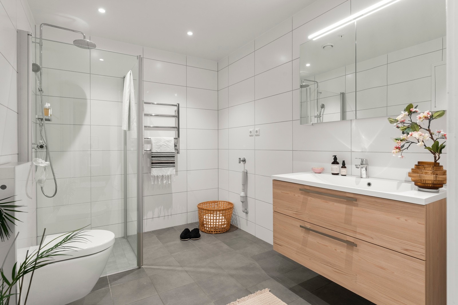 Lekkert og funksjonelt bad med fliser på både gulv og vegger. Rommet har en praktisk kombinasjon av bad og vaskerom, og er innredet med vegghengt toalett, nedsenket servant med stilren innredning, dusjnisje med glassdører og opplegg for vaskemaskin. Galleribilde