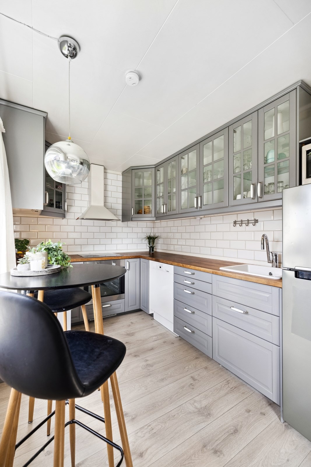 Lyst kjøkken med kjøkkeninnredning i en liten hesteskoformasjon. Innredningen er fra Ikea med profilerte fronter på overskap, underskap og skuffer, samt overskap med glassdører. Galleribilde