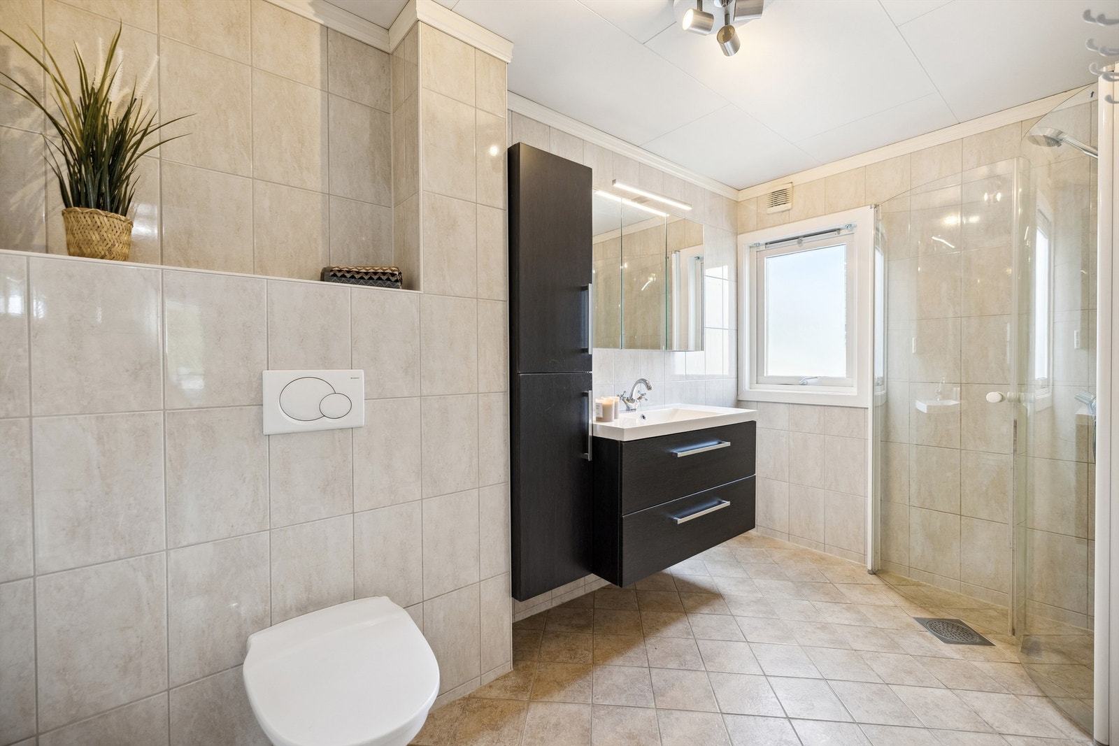 Flislagt baderom med elektriske varmekabler i gulvet. Badet er innredet med heldekkende servant i mørk skuffeinnredning, speilskap, vegghengt høyskap, vegghengt toalett og dusjhjørne med dører og armatur montert på veggen. Galleribilde