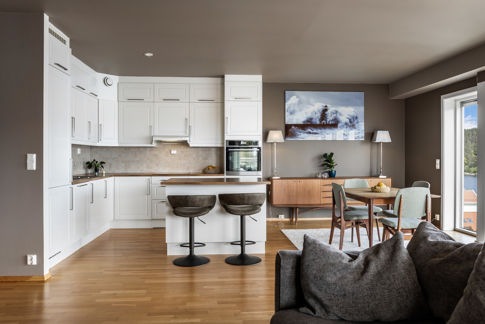 Lyst kjøkken med åpen løsning til stue, spiseplass og store vindusflater som gir godt med naturlig lys. Galleribilde