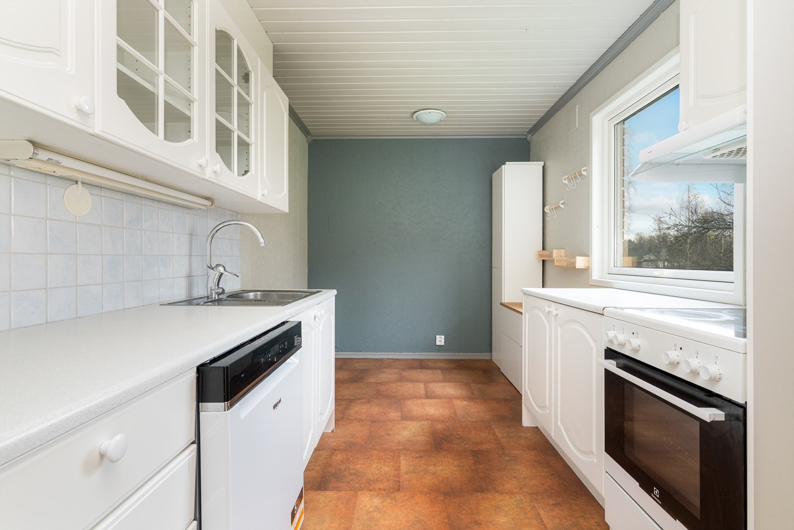 Lyst kjøkken med eldre kjøkkeninnredning. Innredningen har profilerte fronter og laminat benkeplate med nedfelt benkebeslag i rustfritt stål. Galleribilde