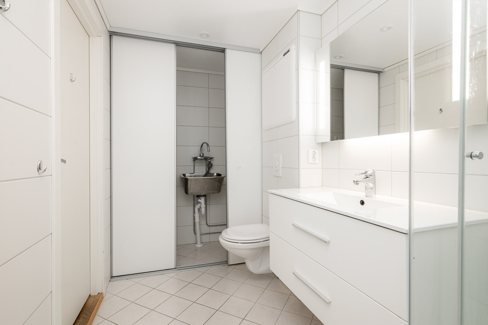 Bred servantinnredning og stort speil med integrert belysning. Skyvedører til vaskeromsdel utstyrt med utslagsvask og opplegg for vaskemaskin. Galleribilde