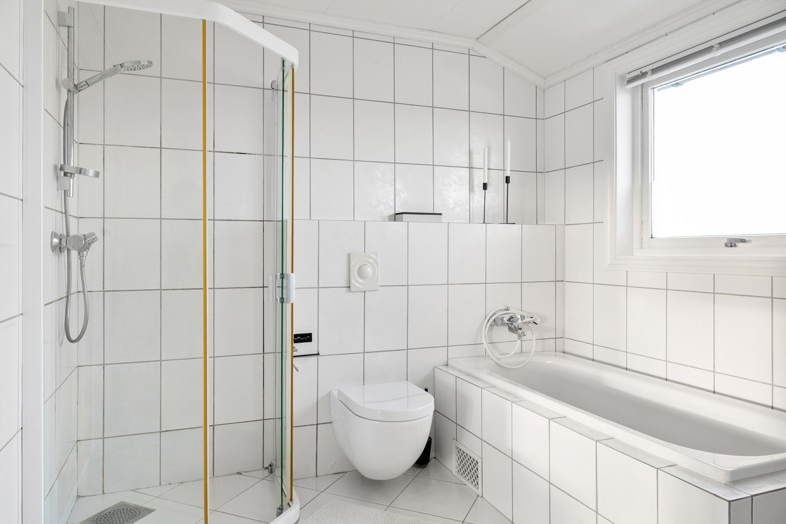 Et flislagt, tidløst bad innredet med dusjhjørne, vegghengt toalett, badekar og servantseksjon. Galleribilde