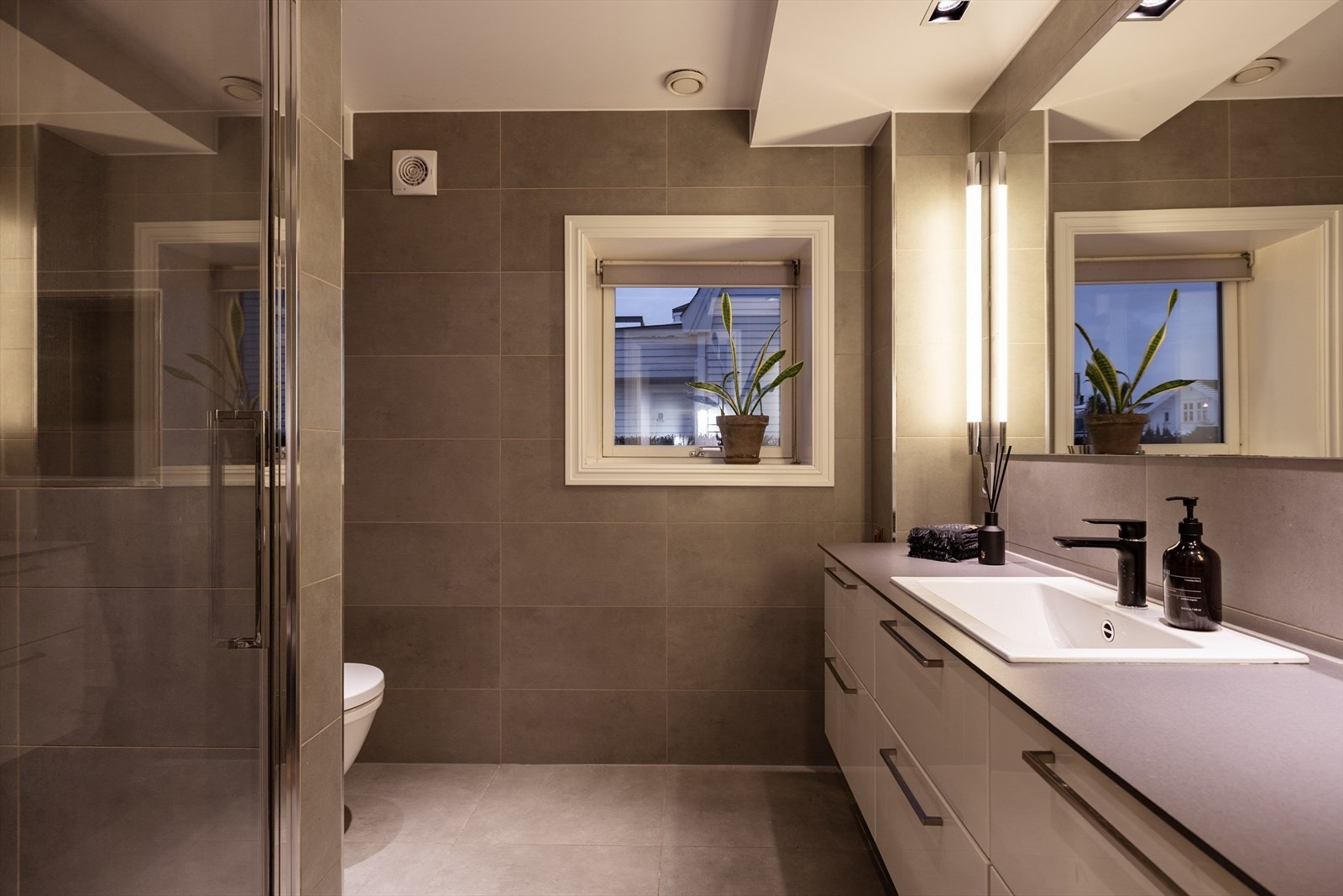 Badet i første etasje fremstår som stilrent og praktisk. Rommet har grå fliser på gulv og vegger, vegghengt toalett, moderne baderomsinnredning med høyglansfronter, servant med speil og belysning, samt en dusjsone. Galleribilde