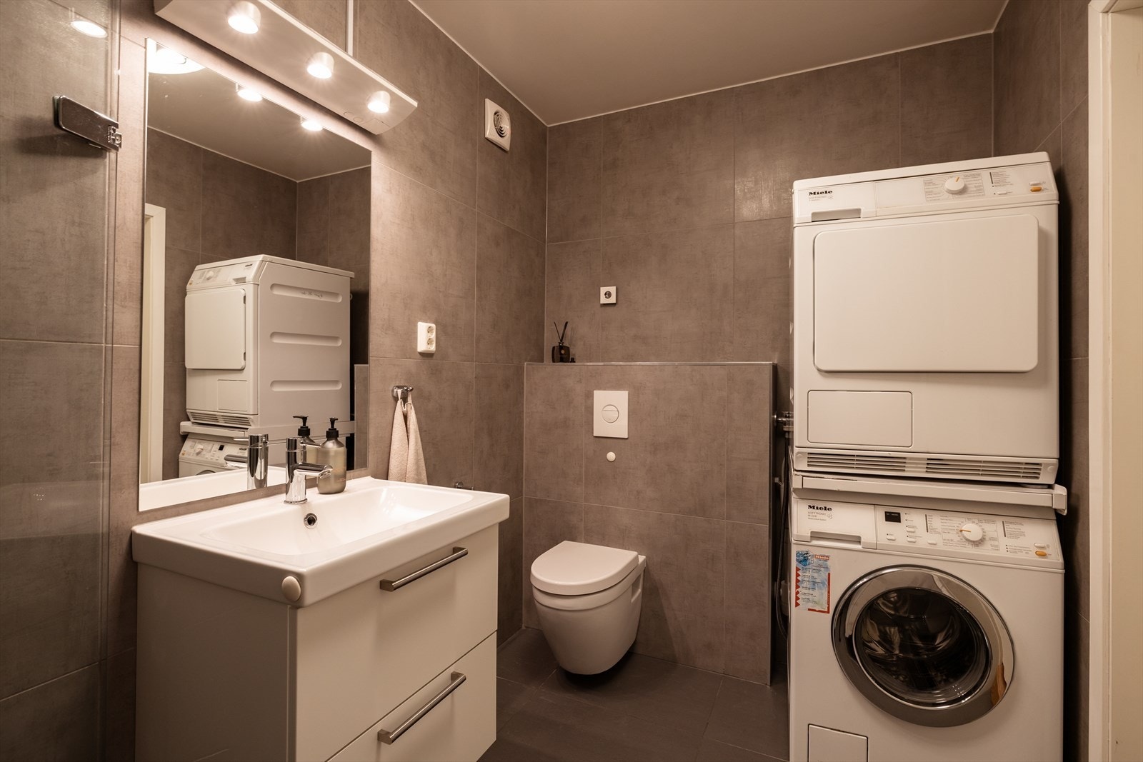 Badet i hybelen er utstyrt med flislagte overflater, moderne baderomsinnredning med glatte fronter, dusj med innfellbare glassdører, servant og vegghengt toalett med innebygd sisterne. Det er også opplegg for vaskemaskin. Galleribilde