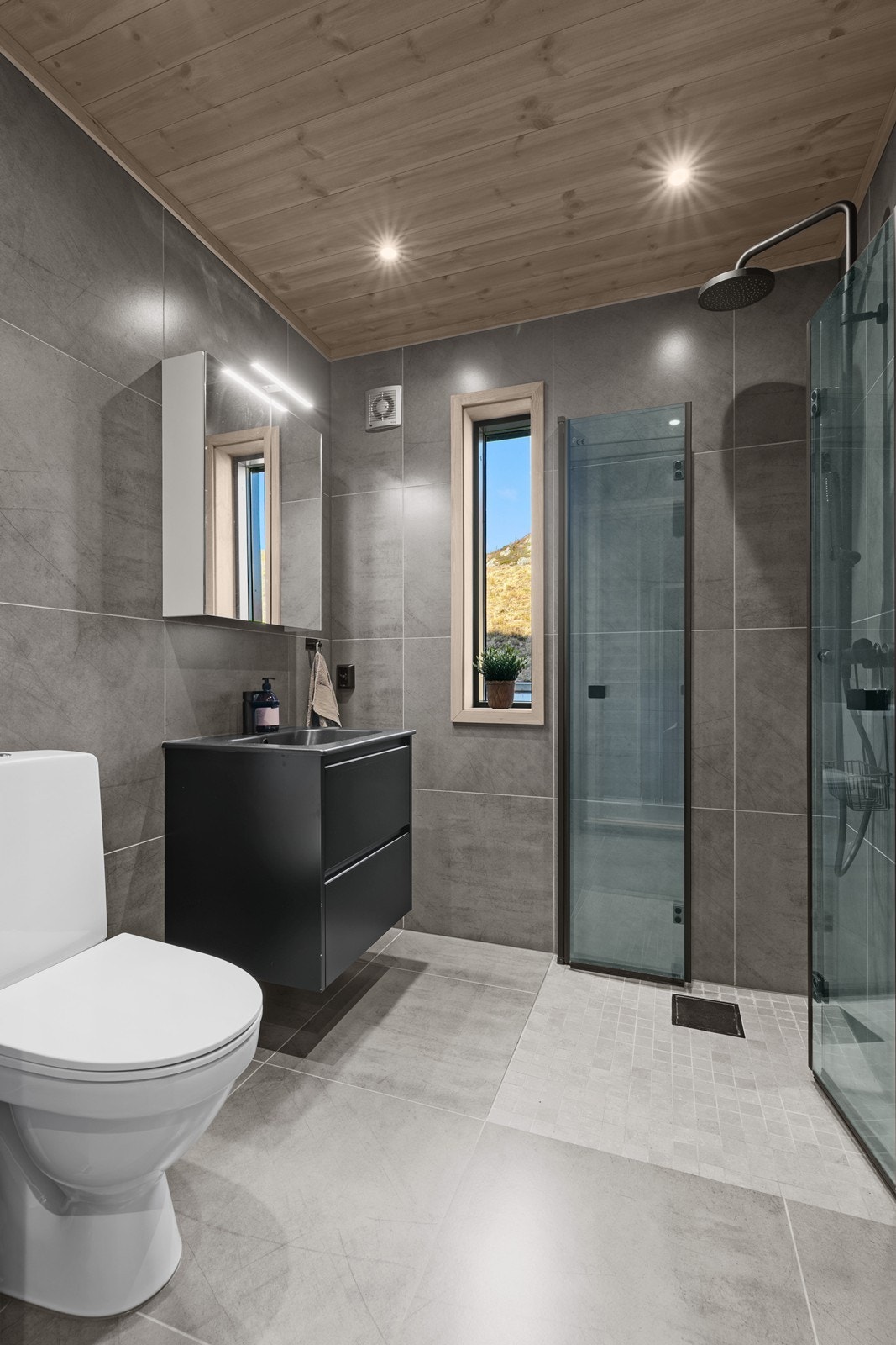 Badet er i gjennomført stil med flislagt gulv og vegger, gulvvarme, dusjnisje med glassdører og regndusj, gulvmontert toalett og heldekkende servant med ettgreps blandebatteri. Galleribilde