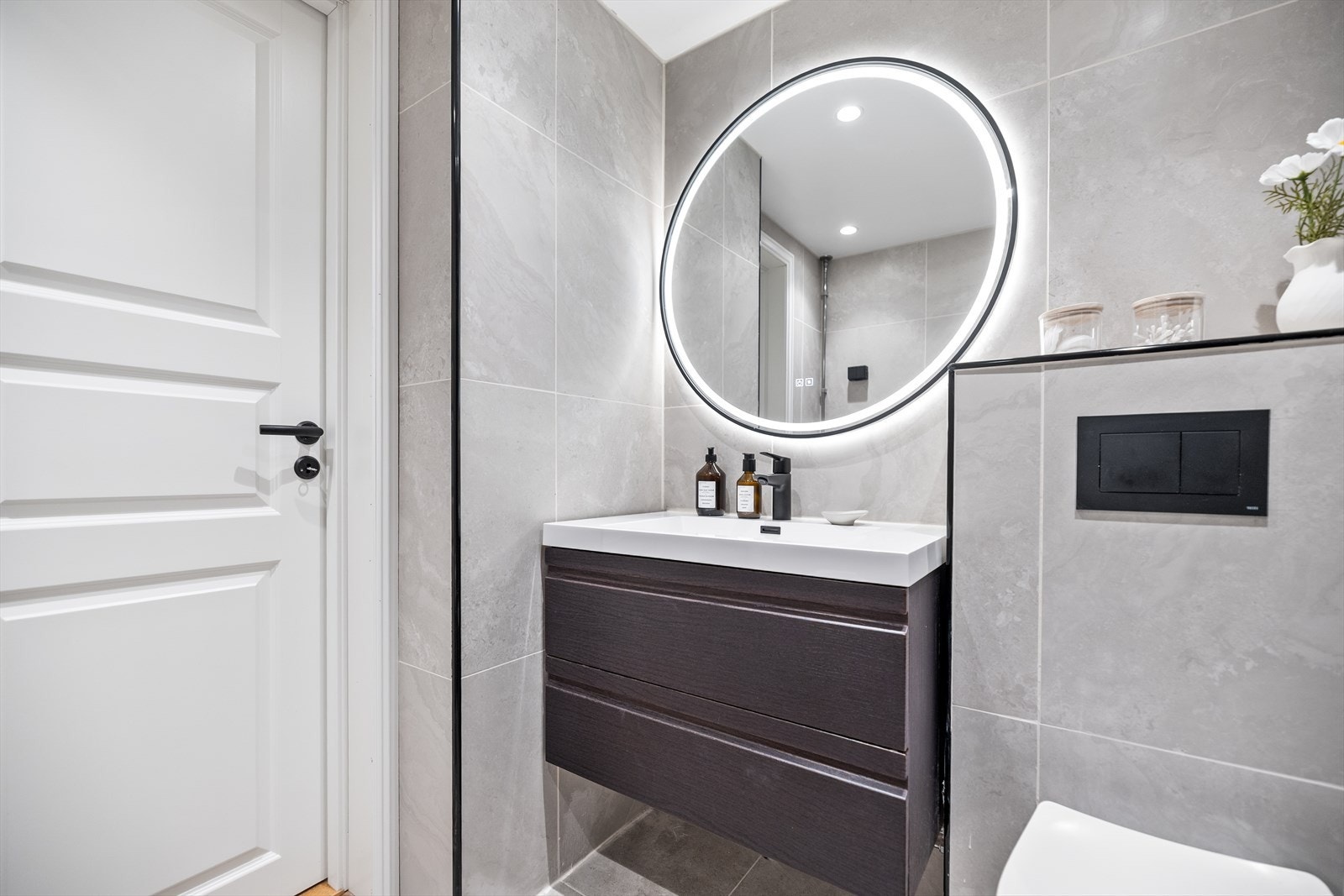 mørke baderomsinnredningen skaper en elegant helhet, mens det runde speilet med integrert belysning tilfører både funksjonalitet og en behagelig atmosfære. Galleribilde