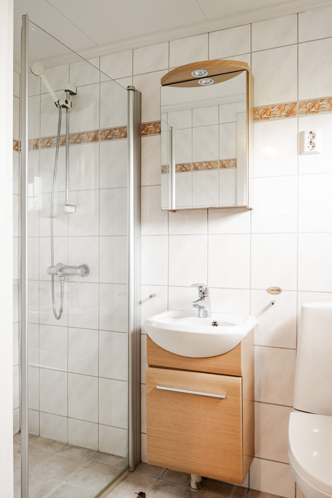 Badet i første etasje har flislagte gulv og vegger, og varmes opp ved varmekabler i gulv. Rommet er innredet med dusjhjørne, servant på benkeskap, frittstående toalett og belyst speil. Galleribilde