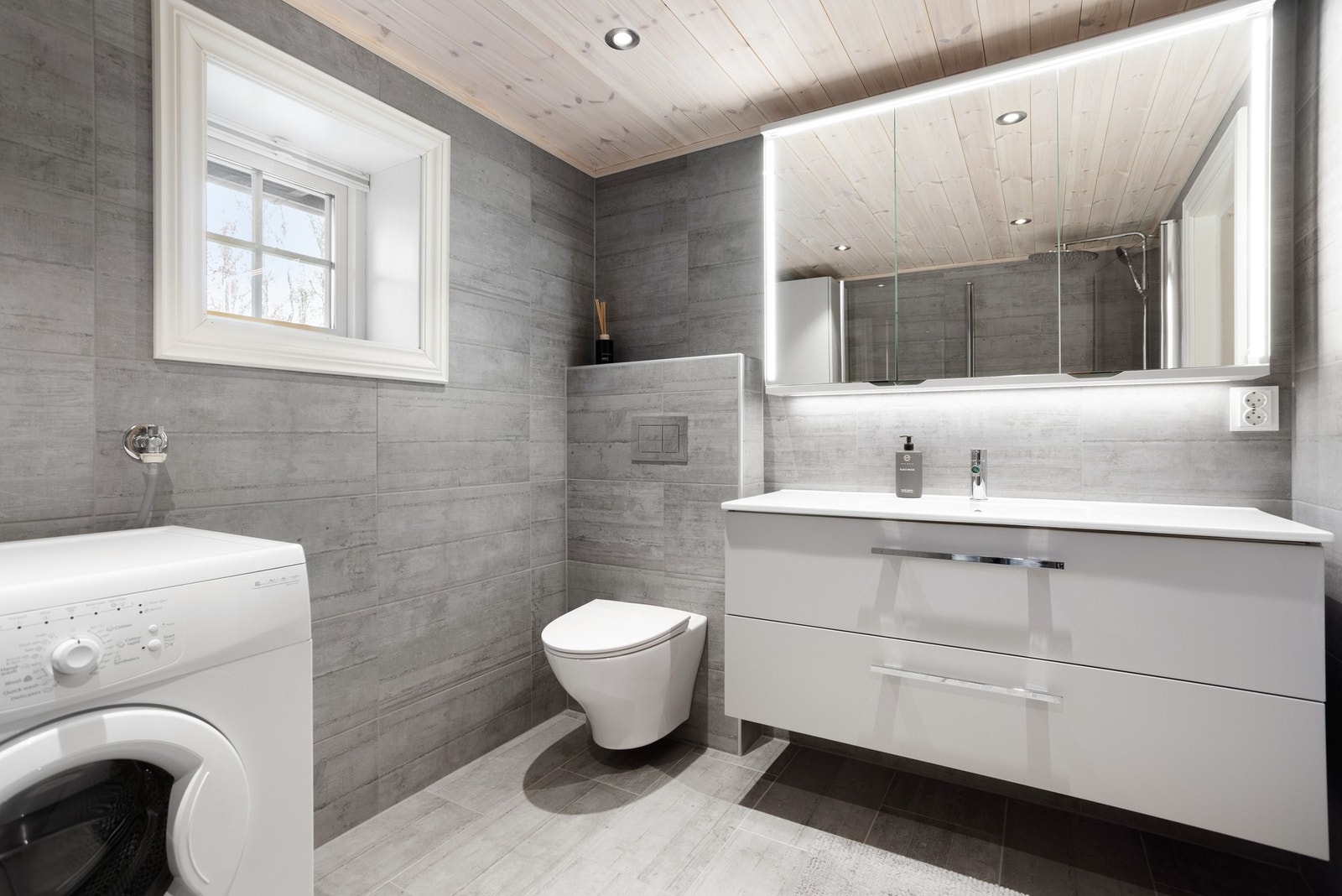 Moderne, flislagt baderom med vegghengt toalett, innredning med servant, dusjhjørne og opplegg til vaskemaskin Galleribilde