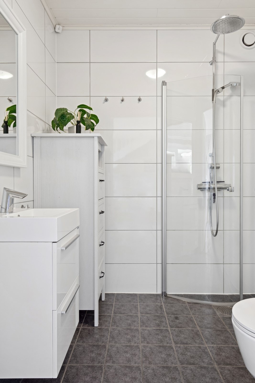 Hvit glatt, flislagt baderom med gulvvarme. Inneholder servant i baderomsinnredning, vegghengt toalett og dusjhjørne med innfellbare glassdører. Galleribilde