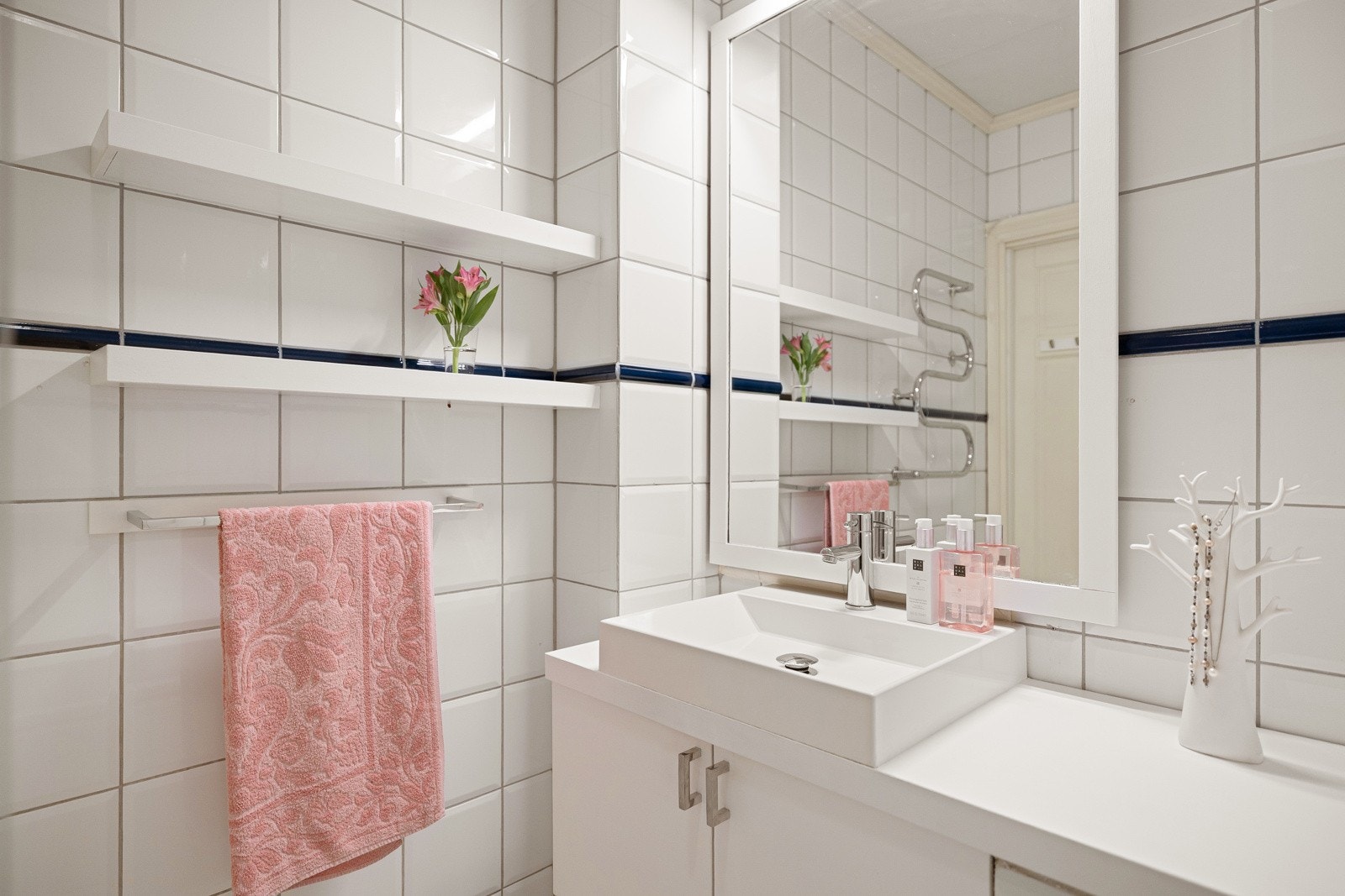 Lys baderomsinnredning med benkeplate og ovenpåliggende servant, speil med belysning over, samt vegghengte hyller ved siden av. Galleribilde