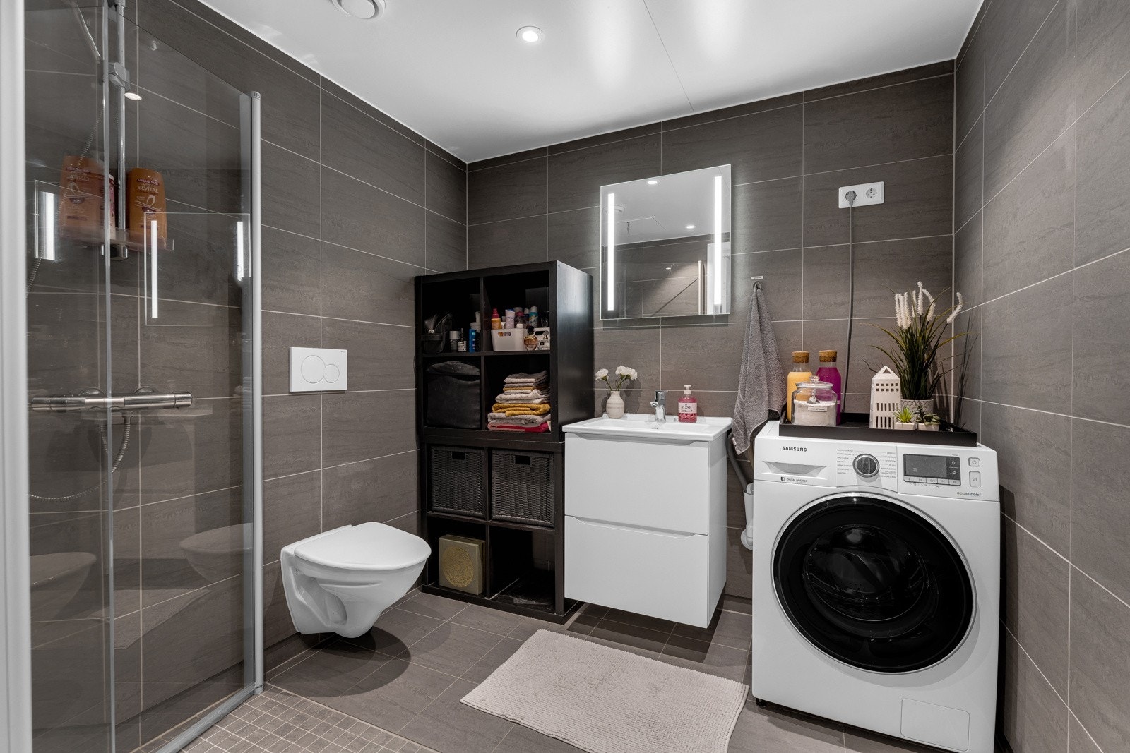 Moderne baderomsinnredning med skuffer og heldekkende servant, speil med integrert belysning over. I hjørnet er det plass og opplegg for vaskemaskin, samt tørketrommel over om ønskelig. Galleribilde